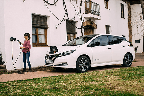 Eine Frau lädt ihren weißen, vollelektrischen Nissan Leaf im Vorgarten über eine Wallbox an der Hauswand auf.